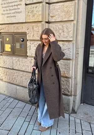 Mujer con abrigo largo marrón, jeans anchos azul claro y zapatos beige, sosteniendo un bolso negro, de pie junto a una pared de piedra con texto en alemán.