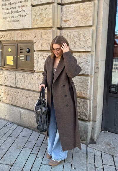Mujer con abrigo largo marrón, jeans anchos azul claro y zapatos beige, sosteniendo un bolso negro, de pie junto a una pared de piedra con texto en alemán.