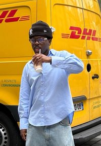 Person som holder en drink, iført en lyseblå skjorte med knapper og falmede denimbukser, med en svart lue, foran en gul DHL-varebil.