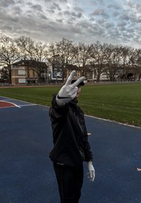 Fekete dzseki és fekete nadrág; szürke sportkesztyűk logóval. Egy személy békejelet mutat egy kék pályán, háttérben zöld fű és fák.