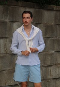 Camisa de botão azul clara com riscas, suéter branco colocado sobre os ombros, calções azul claros, de pé contra uma parede cinza texturizada.