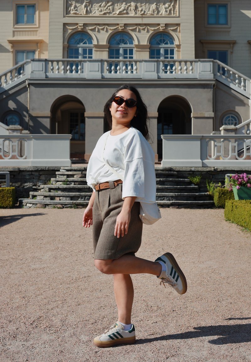 Kvinde med solbriller poser på et ben iført hvid oversized top, brune shorts og sneakers med historisk bygningsfacade i baggrunden.