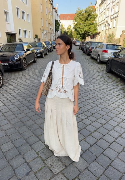 Top de encaje blanco con diseño recortado, atado al cuello; combinado con una falda beige de tiro alto y pliegues. Escena de calle con coches aparcados.