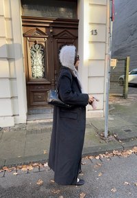 Lang sort frakke med dobbeltradet design, grå hætte foret med pels og sort taske; kombineret med sorte sko, stående på en fortov.