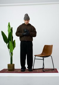 Brązowa bluza zapinana na zamek z kapturem, czarne rękawice, szerokie czarne spodnie, czarne obuwie, obok zielonej rośliny w prążkowanej doniczce i brązowego krzesła.