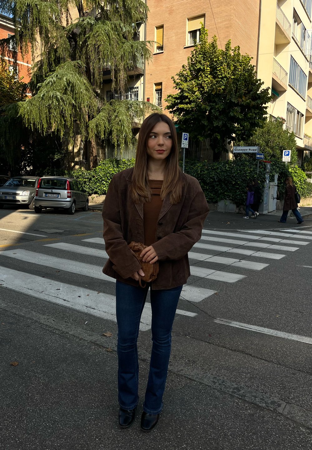 Brown suede jacket, brown top, and flared blue jeans. Standing on a crosswalk with a small brown bag, surrounded by trees and buildings.