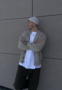 Cardigan beige tricoté avec boutons, porté sur un t-shirt blanc. Pantalon foncé et bonnet gris clair. Appuyé contre un mur lisse gris.