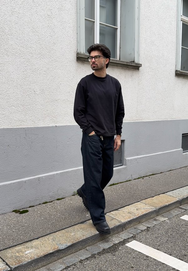 Άνδρας με σκούρα μαλλιά και γυαλιά, φοράει μαύρο φούτερ και φαρδιά μαύρη παντελόνια, περπατά σε ένα γκρι πεζοδρόμιο δίπλα σε ένα λευκό κτίριο.