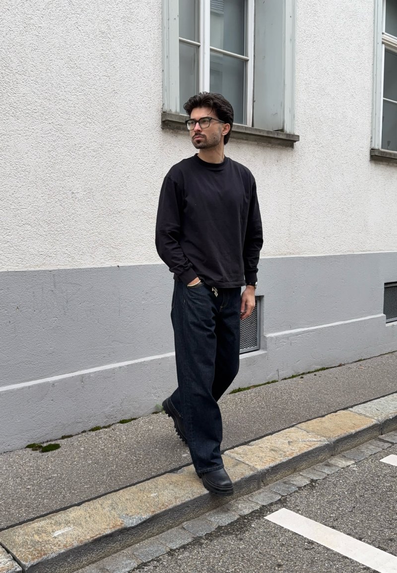 Muž s tmavými vlasmi a okuliarmi, oblečený v čiernej mikine a širokých čiernych nohaviciach, kráča po šedom chodníku vedľa bielej budovy.