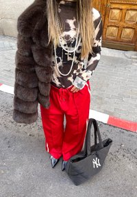 Top nero trasparente con motivo astratto, pantaloni da jogging rossi, cappotto in pelliccia marrone, collane argentate e una borsa a tracolla nera trapuntata con logo bianco. Scarpe nere a punta.