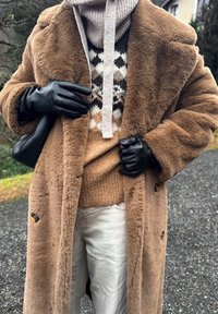 Άτομο που φοράει καφέ παλτό από συνθετική γούνα, πουλόβερ με σχέδια, μπεζ παντελόνι, μαύρα δερμάτινα γάντια και κρατά μια μαύρη δερμάτινη τσάντα σε εξωτερικό χώρο.