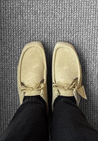 Beżowe mokasyny z zamszu w stylu mokasynowym, z zaokrąglonym noskiem, sznurowane cienkimi sznurkami, na tle szarego teksturowanego dywanu.