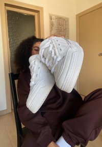 Zapatillas deportivas blancas con upper texturizado y refuerzos en los ojales. Suelas blancas gruesas, punteras redondeadas y cordones. Conjunto casual de fondo.