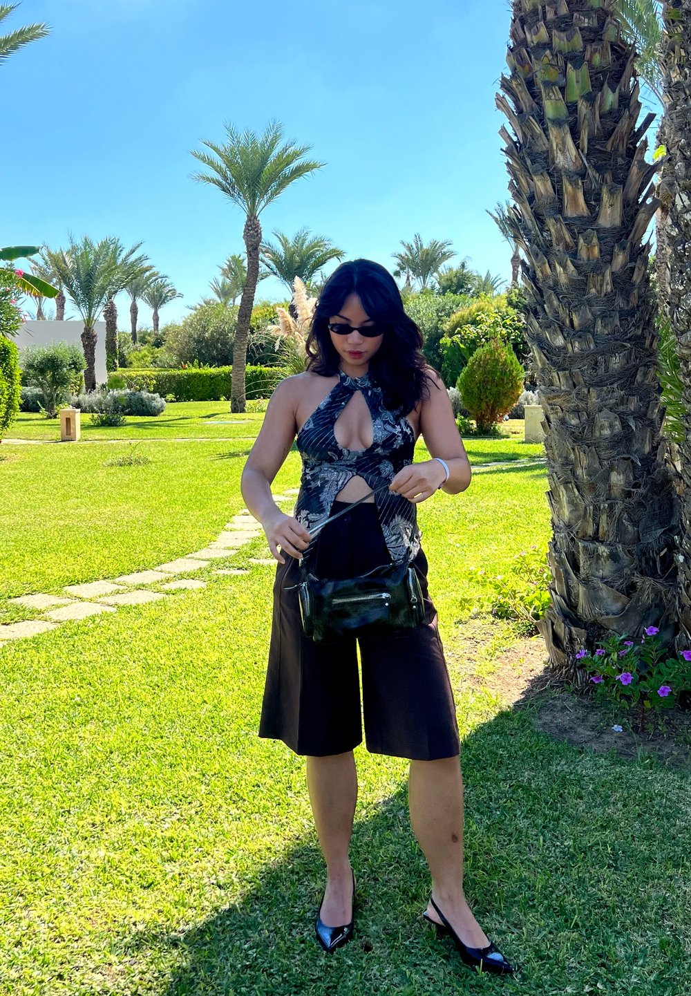 Top a fascia con motivo floreale, pantaloni corti larghi neri, borsa nera e occhiali da sole. Erba verde e palme sullo sfondo.
