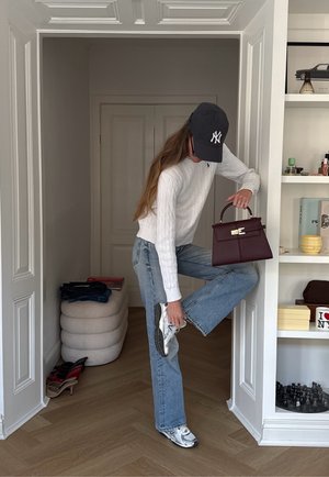 Persona con suéter blanco, jeans azules y gorra negra atándose los tenis mientras sostiene un bolso burdeos en una habitación minimalista.