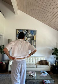 Λευκό μπλουζάκι και γκρι φόρμες φορεμένα από μια φιγούρα, στραμμένη προς ένα μοντέρνο σαλόνι με έναν καναπέ σε κρέμ χρώμα και μ�ια γυάλινη τραπεζαρία.