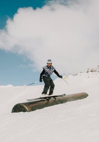 Esquiador con ropa negra y camuflaje deslizándose sobre un riel metálico en una pendiente nevada bajo un cielo azul parcialmente nublado.