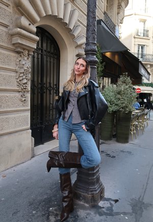 Mujer con chaqueta de cuero negra, camiseta de rayas, vaqueros azules y botas marrones apoyada en un farola en una acera de la ciudad.