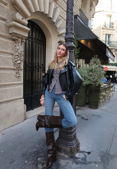 Mujer con chaqueta de cuero negra, camiseta de rayas, vaqueros azules y botas marrones apoyada en un farola en una acera de la ciudad.