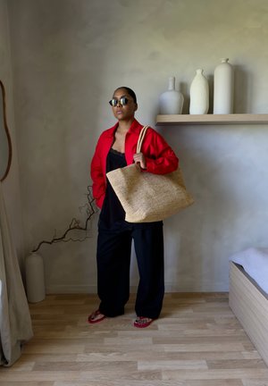 Mujer con chaqueta roja, pantalones negros y gafas de sol sostiene una gran bolsa tejida mientras está de pie en una habitación minimalista con jarrones en la estantería y suelo de madera.