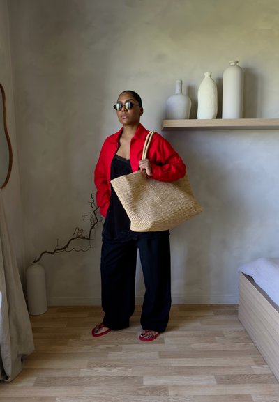 Mujer con chaqueta roja, pantalones negros y gafas de sol sostiene una gran bolsa tejida mientras está de pie en una habitación minimalista con jarrones en la estantería y suelo de madera.