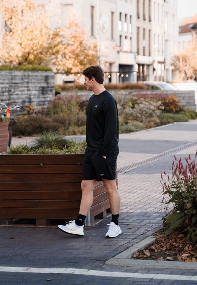 Camisa negra de manga larga y pantalones cortos negros que lleva un hombre caminando por un camino pavimentado, con vegetación y jardineras de madera de fondo.