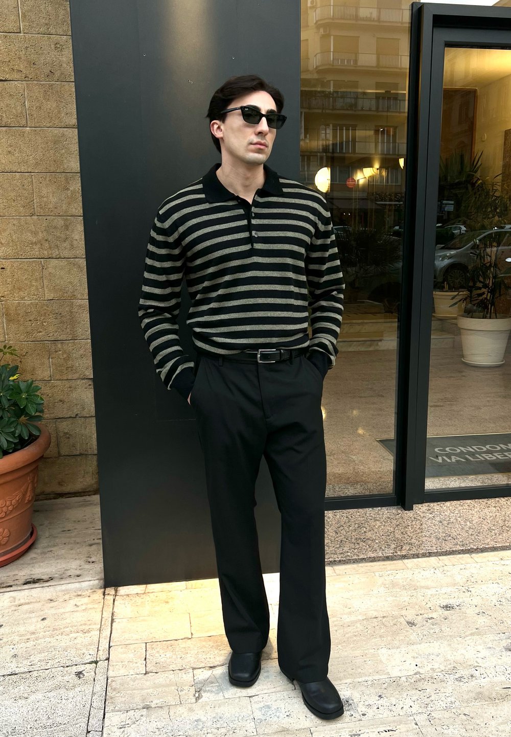 Polo shirt a righe nere e oliva, pantaloni neri e scarpe nere. In piedi in una porta con piante in vaso e una facciata di vetro sullo sfondo.