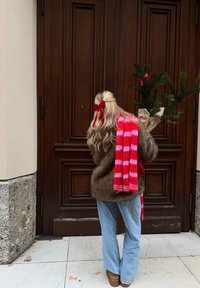 Person with long hair wearing a brown fur coat and light blue jeans, carrying a small evergreen tree wrapped in paper, red tartan scarf draped over shoulder.