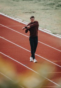 Un athlète portant un haut à manches longues marron et un pantalon noir s'étire sur une piste rouge. Il porte des chaussures de course blanches, avec de l'herbe verte en arrière-plan.
