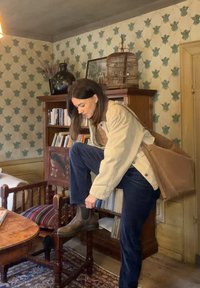 Beige jas, marineblauwe broek en bruine chelsea boots. Houten stoel met patroon op de zitting, boekenplank en vintage decor in een zwak verlichte kamer.