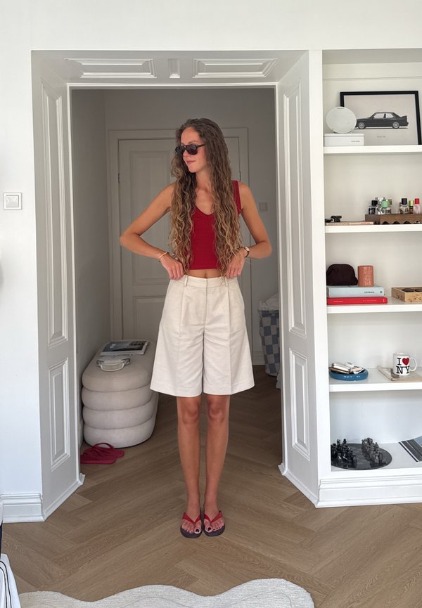 Vrouw met lang krullend haar, rood hemdje, beige korte broek en zonnebril die haar tailleband verstelt in een witte kamer met planken en een poef.