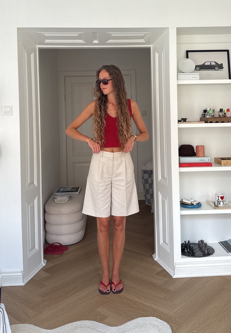 Kvinne med langt krøllete hår, rød singlet, beige shorts og solbriller som justerer livvidden i et hvitt rom med hyller og ottoman.