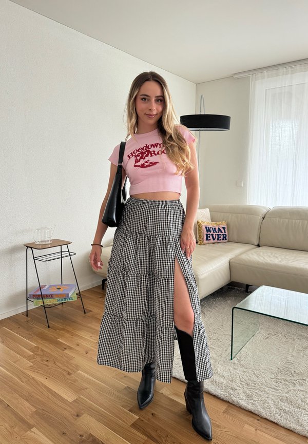 Top cropped rosa com gráfico vermelho; saia maxi de gingle preta e branca com fenda lateral; botas pretas de tornozelo. Fundo simples com mobiliário.