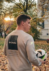 Μπεζ πουκάμισο με μακριά μανίκια και μαύρο λογότυπο Columbia στην πλάτη. Το άτομο κρατά μια υφασμάτινη ποδοσφαιρική μπάλα με διακοσμητική επιφάνεια, περιτριγυρισμένο από φύλλα φθινοπώρου.