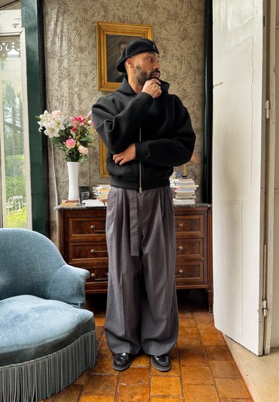 Sudadera negra con cremallera, pantalones anchos grises y zapatos negros. Entorno interior con una silla azul, un arreglo floral y un armario de madera.