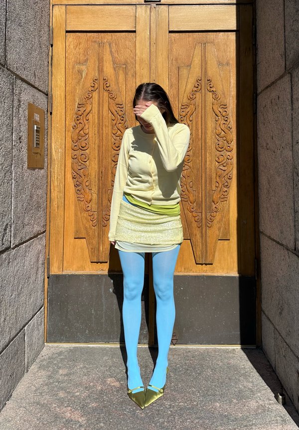 Kobieta w żółtym topie, jasnozielonej mini spódnicy, niebieskich rajstopach i zielonych szpilkach osłaniająca oczy przed światłem słonecznym, stojąca przed ozdobionymi drewnianymi drzwiami.