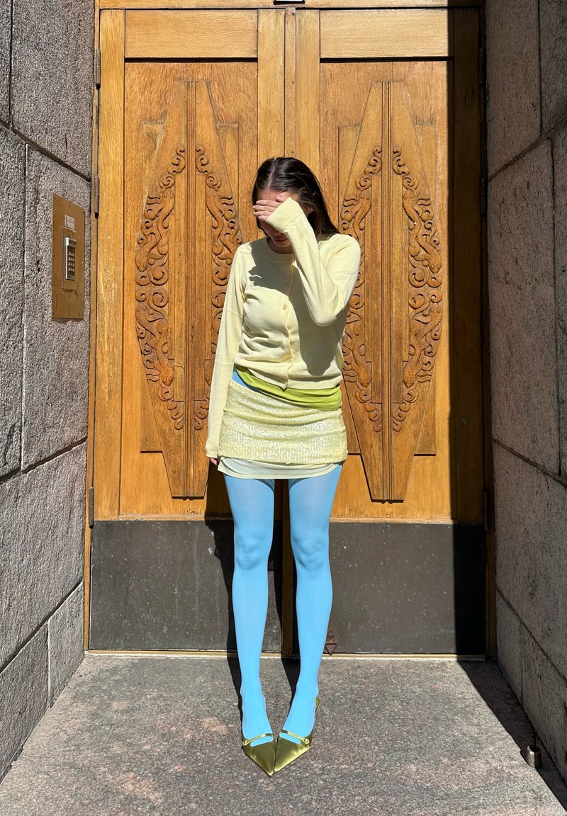 Frau in gelbem Top, hellgrünem Minirock, blauen Strumpfhosen und grünen Absätzen hält sich die Augen vor Sonnenlicht geschützt und steht vor einer verzierten Holztür.