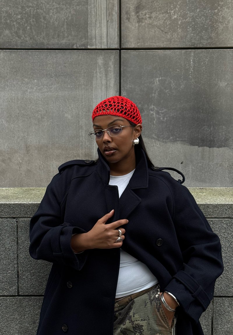 Musta, kaksirivinen takki leveillä kauluksilla, punainen virkattu huivi päässä ja kirkkaat silmälasit, harmaata tekstuuriseinää vasten.