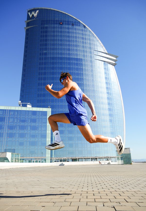 Hombre con ropa deportiva azul y gafas de sol saltando frente a un alto edificio de vidrio curvo bajo un cielo azul despejado.