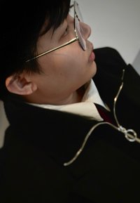 Jeune personne portant des lunettes à monture dorée, chemise blanche à col et veste noire avec une tirette circulaire, vue de profil de près, face à droite.