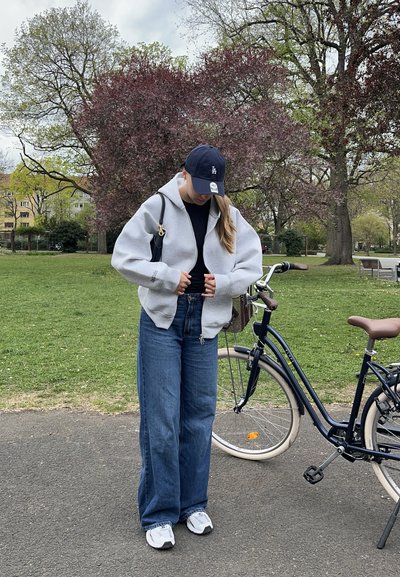 Sudadera gris con cremallera, camiseta negra, jeans anchos azules, zapatillas blancas. Lleva una gorra oscura y sostiene un bolso. Bicicleta clásica cercana en la acera.