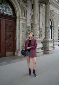 Υπεργαλαντωμένο μάρκετ μπουφάν σε μπορντό απόχρωση πάνω από μαύρη μπλούζα, γκρίζα μίνι φούστα, μπορντό κάλτσες και μεταλλικά τακούνια, κρατώντας μια μαύρη τσάντα μπροστά από μια περίτεχνη οικοδόμηση.