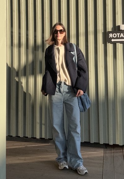 Abrigo de gran tamaño en color navy, suéter crema, jeans azules claros y zapatillas blancas. Bolso de hombro de tela de mezclilla. De pie contra una pared verde.
