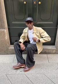 Casaco bege sobre uma t-shirt branca, calças pretas largas e sapatos castanhos. A pessoa está sentada no chão ao lado de uma porta verde escura.