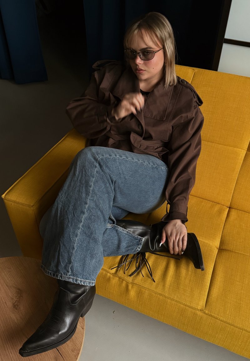 Jeune femme portant des lunettes de soleil, une veste marron, un jean et des bottes noires, assise jambes croisées sur un canapé jaune dans une pièce faiblement éclairée.