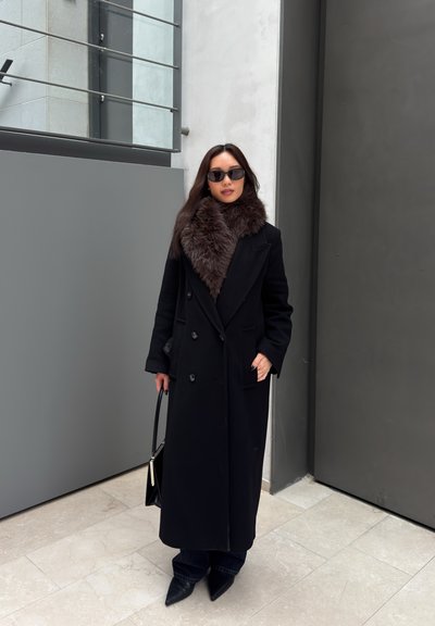 Mujer de pie en el interior, lleva un abrigo largo negro con cuello de piel, gafas de sol negras, botas puntiagudas y sostiene un bolso negro.