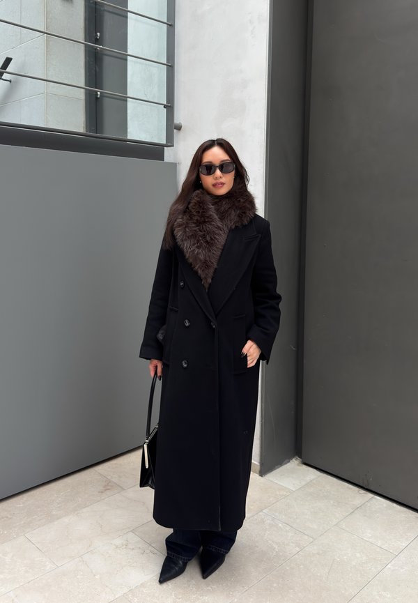 Žena stojaca vo vnútri, oblečená v dlhom čiernom kabáte s kožušinovým golierom, čiernych slnečných okuliaroch, špicatých čižmách a držiaca čiernu kabelku.