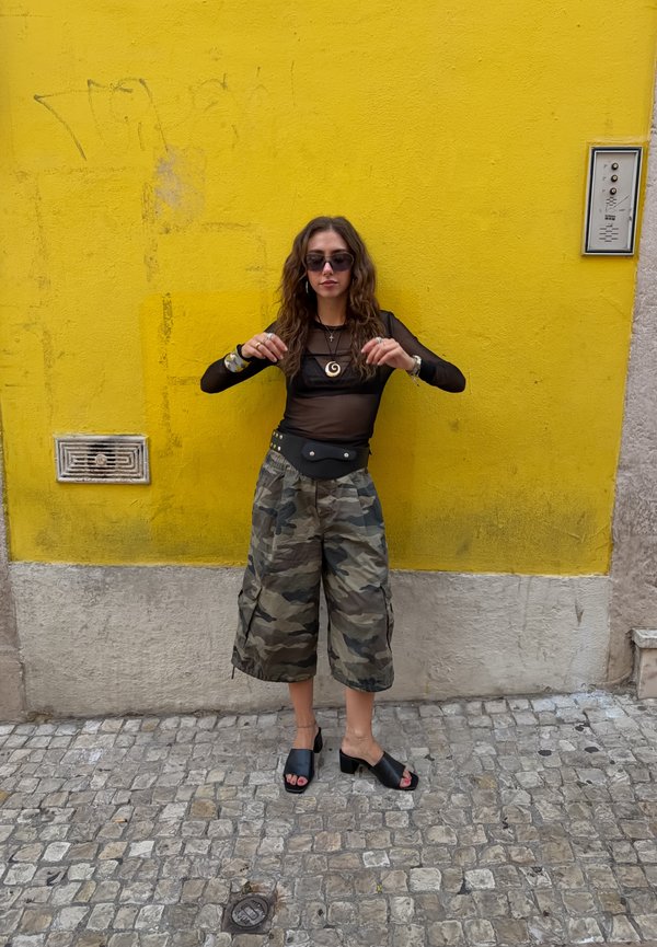 Frau mit Sonnenbrille trägt schwarzen transparenten Oberteil, weite Camouflage-Hosen und schwarze Sandalen mit Absatz, steht auf Kopfsteinpflaster vor einer gelben Wand.