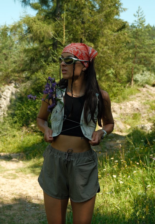 Mujer joven con gafas de sol, bandana roja y ropa de exterior sosteniendo flores moradas en un claro soleado del bosque.