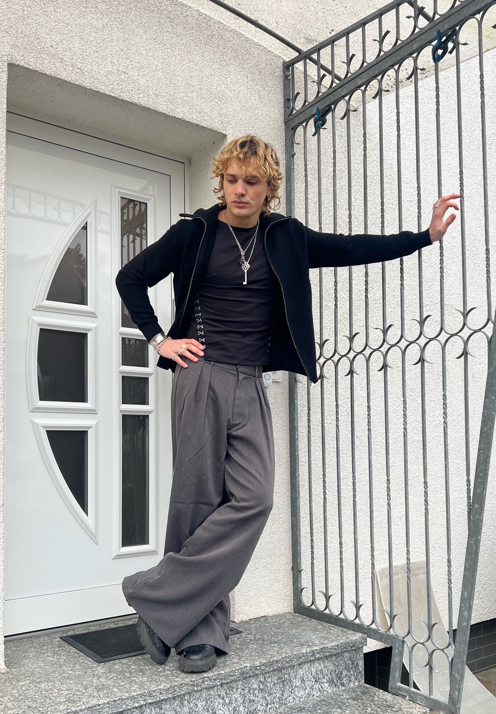 Jeune personne aux cheveux bouclés appuyée contre une grille métallique, portant un pantalon large gris, une chemise noire, une veste noire et des chaussures noires épaisses sur des marches en pierre.